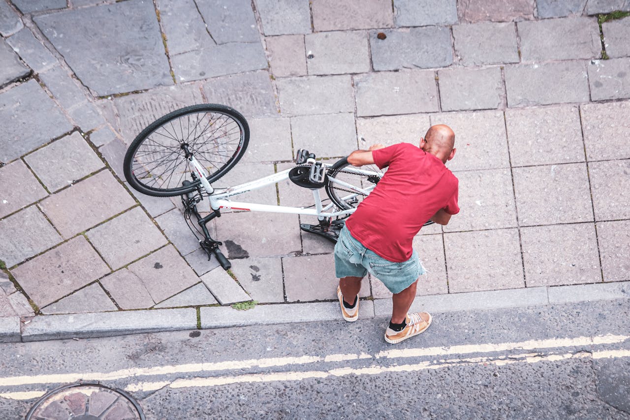 A bald man in a red shirt fixing a bike on a city sidewalk. High angle view.